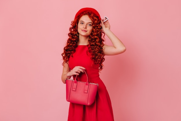 Portret ładnej pani z falowanymi rudymi włosami i niebieskimi oczami. Dziewczyna w czerwonej sukience wkłada beret i trzyma jej torbę.