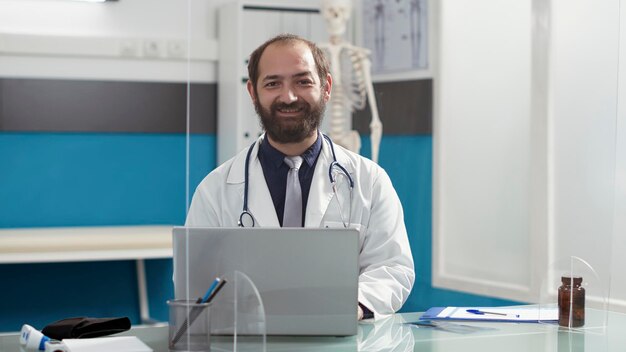 Portret lekarza rodzinnego pracującego na laptopie przy biurku, korzystającego z komputera do udzielania wsparcia pacjentom podczas wizyty kontrolnej. Mężczyzna lekarz ubrany w biały fartuch i stetoskop.