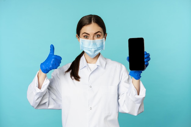 Portret lekarza w medycznej masce na twarz i rękawiczkach pokazujący ekran aplikacji telefonu komórkowego i smartfona i t...