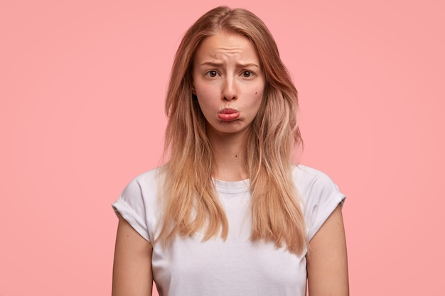 Bezpłatne zdjęcie portret maltretowanej blondynki dąsa się i zaciska usta, jest obrażana przez kogoś i czeka na przeprosiny, ma proste włosy, odizolowane na różowej ścianie