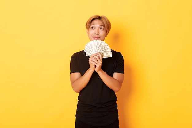 Bezpłatne zdjęcie portret marzycielskiego przystojnego azjata pokazującego swoje oszczędności i myślącego, patrząc w lewym górnym rogu, trzymając pieniądze, stojąc na żółtej ścianie