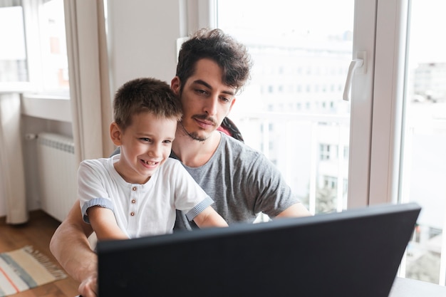 Bezpłatne zdjęcie portret mężczyzna obsiadanie z jego uśmiechniętym synem używa laptop w domu