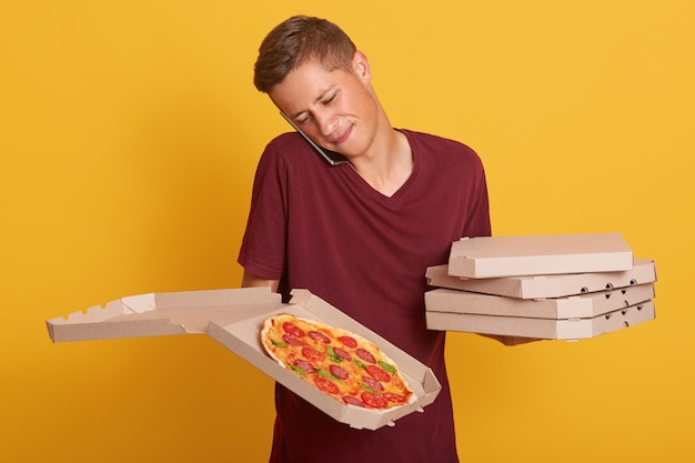 Bezpłatne zdjęcie portret mężczyzny mówiącego przez telefon, noszącego burgundową swobodną koszulkę, trzymającego pudełka z pizzą, otrzymuje nowe zamówienie za pośrednictwem swojego smartfona