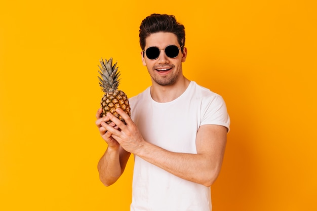 Portret mężczyzny w białej koszulce i okularach przeciwsłonecznych, trzymając ananas na pomarańczowej przestrzeni.