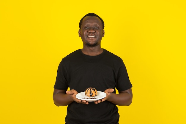 Portret mężczyzny w czarnej koszulce trzymającego talerz ciasta z polewą czekoladową na tle żółtej ściany