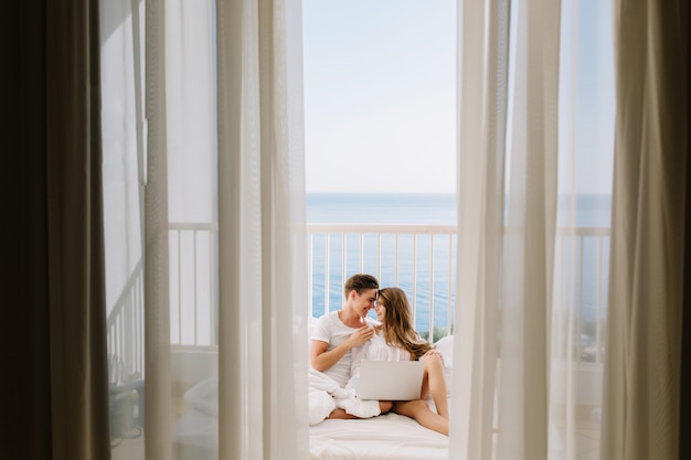 Portret miłości para razem oglądanie filmu na balkonie rano z zasłonami na pierwszym planie. Młody człowiek odpoczywa ze swoją niesamowitą opaloną dziewczyną w łóżku trzymając laptopa z widokiem na morze