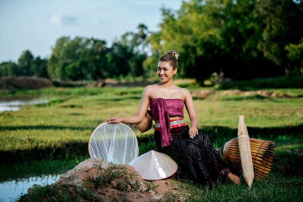Portret Młoda ładna Azjatycka kobieta w pięknych tajskich tradycyjnych strojach na polu ryżowym, siedzi w pobliżu sprzętu wędkarskiego