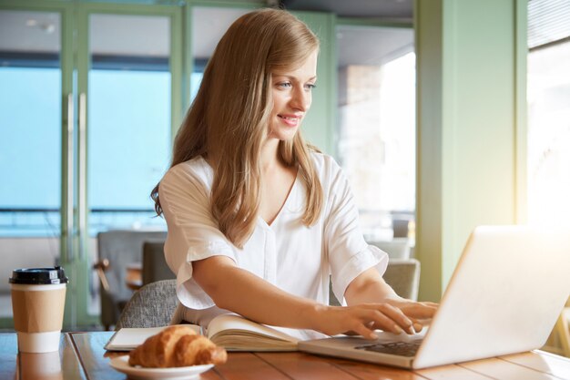 Portret młoda zrelaksowana kobieta pisać na maszynie na laptopu sittingat cukiernianego stół