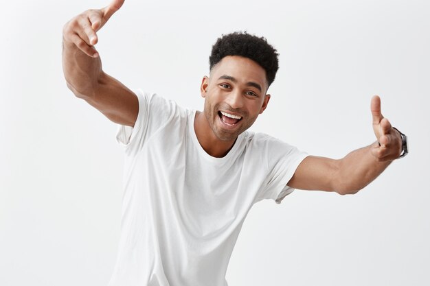 Portret młodego atrakcyjnego, ciemnoskórego mężczyzny z fryzurą afro w białej swobodnej koszulce uśmiechniętej zębami, gestykulując rękami w aparacie, patrząc w kamerę z radosnym i podekscytowanym wyrazem twarzy