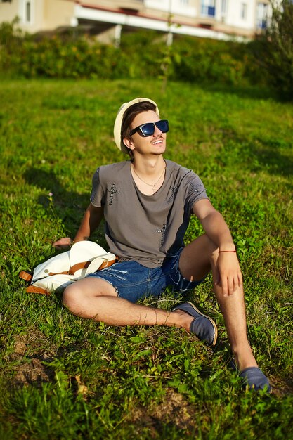 portret młodego atrakcyjnego nowoczesnego stylowego mężczyzny w dorywczo tkaniny w kapeluszu w okularach siedzi w parku w zielonej trawie