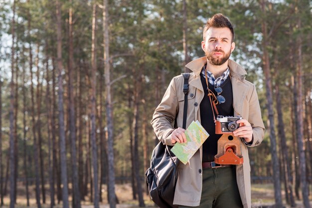 Portret młodego człowieka stojącego w lesie trzymając mapę i vintage aparatu