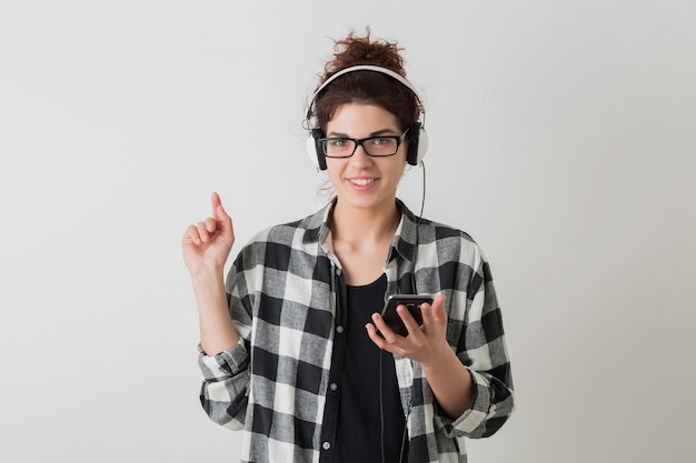 Portret młodego hipster uśmiechnięta ładna kobieta w kraciastej koszuli w okularach pozowanie na białym tle, trzymając inteligentny telefon i słuchanie muzyki w słuchawkach