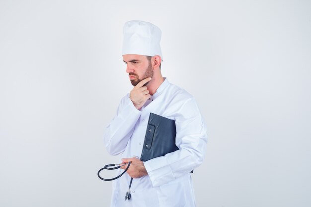 Portret młodego lekarza płci męskiej trzymając schowek, stetoskop, dotykając brodę w białym mundurze i patrząc zamyślony widok z przodu