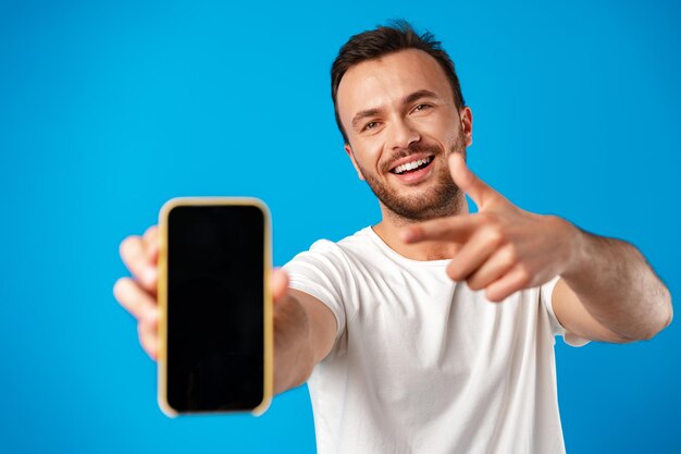Portret młodego mężczyzny reklamującego nowy smartfon pokazujący go kamerze na niebieskim tle