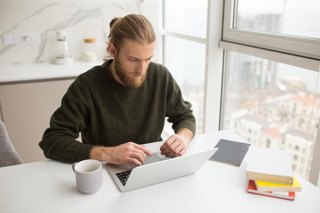 Portret młodego mężczyzny siedzącego przy stole z kubkiem i książkami podczas pracy na laptopie w domu
