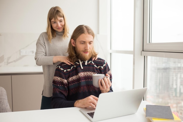 Portret młodego mężczyzny w swetrze, siedzącego przy stole z kubkiem w dłoni i pracującego na laptopie, podczas gdy jego dziewczyna stoi w domu