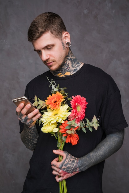 Portret młodego mężczyzny z przebitymi uszami i nosa gospodarstwa kwiat w ręku za pomocą smartfona