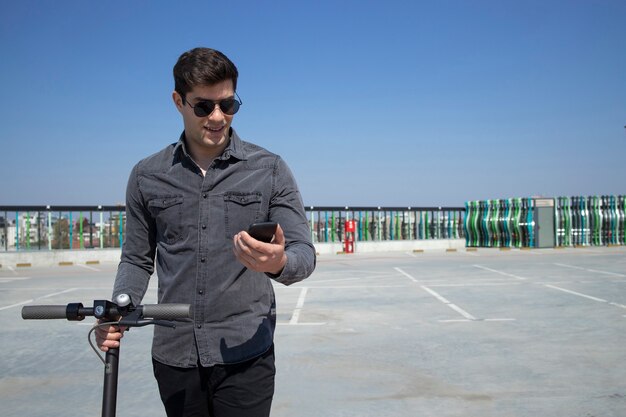 Portret młodego przystojnego mężczyzny stojącego na jego skuter elektryczny i patrząc na jego inteligentny telefon