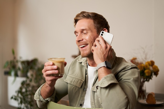 Portret młodego przystojniaka rozmawiającego przez telefon, pijącego kawę i śmiejącego się, facet siedzący w kawiarni.