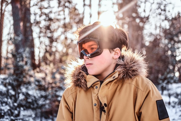 Bezpłatne zdjęcie portret młodego snowboardzisty ubranego w ciepłe ubrania i okulary ochronne, stojącego w zaśnieżonym lesie podczas wschodu słońca