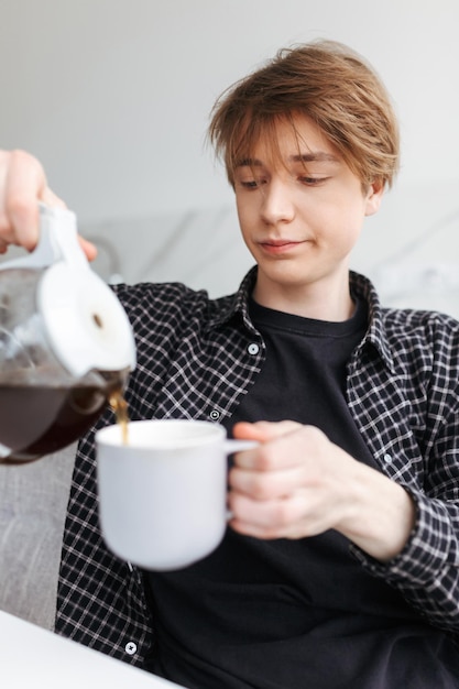 Portret młodego śpiącego mężczyzny siedzącego i nalewającego kawę w kuchni w domu