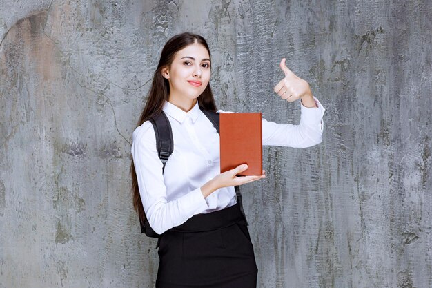 Portret młodego ucznia w stroju szkolnym, trzymając książkę i dając kciuk w górę. Zdjęcie wysokiej jakości