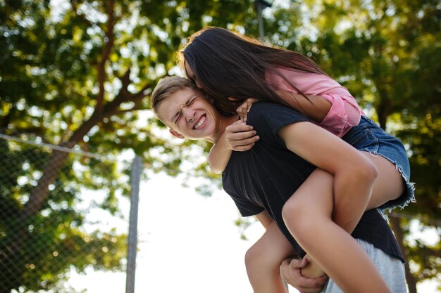 Portret młodego uśmiechniętego chłopca trzymającego ładną dziewczynę na plecach i bawiącego się z nią podczas spędzania czasu w parku