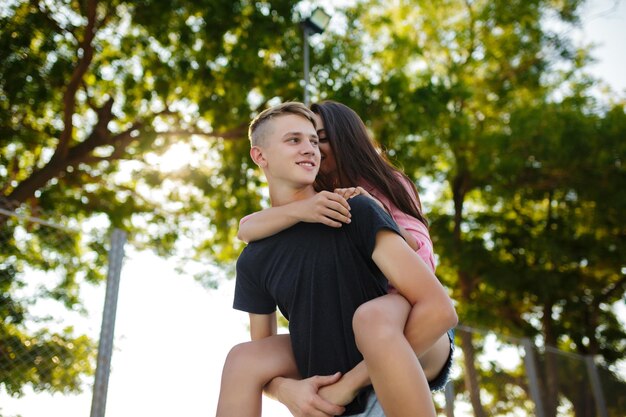 Portret młodego uśmiechniętego chłopca trzymającego ładną dziewczynę na plecach, szczęśliwie patrzącego na bok spędzającego czas w parku