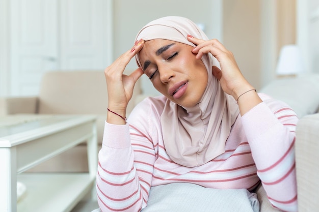 Portret młodej arabskiej muzułmanki siedzącej na kanapie w domu z bólem głowy i bólu kobieta w hidżabie cierpiąca na chroniczne codzienne bóle głowy smutna kobieta trzymająca się za głowę z powodu bólu zatok