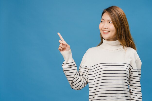 Portret młodej azjatyckiej damy uśmiechający się z radosnym wyrazem twarzy, pokazuje coś niesamowitego w pustej przestrzeni w swobodnym ubraniu i stojącej odizolowanej na niebieskiej ścianie