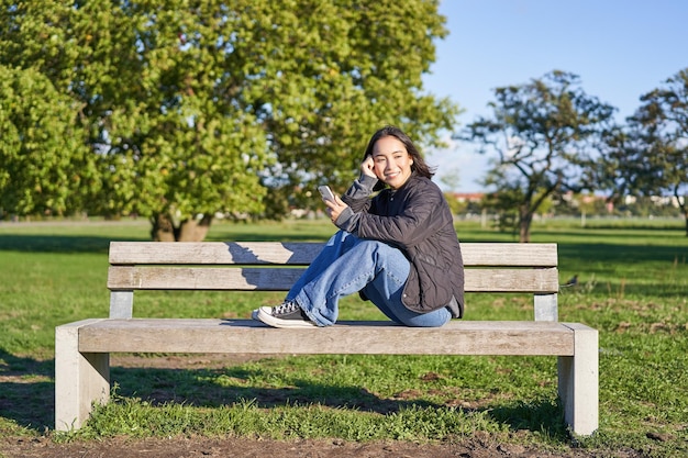 Portret młodej azjatyckiej kobiety siedzącej na ławce ze smartfonem śliczna dziewczyna korzystająca z telefonu komórkowego