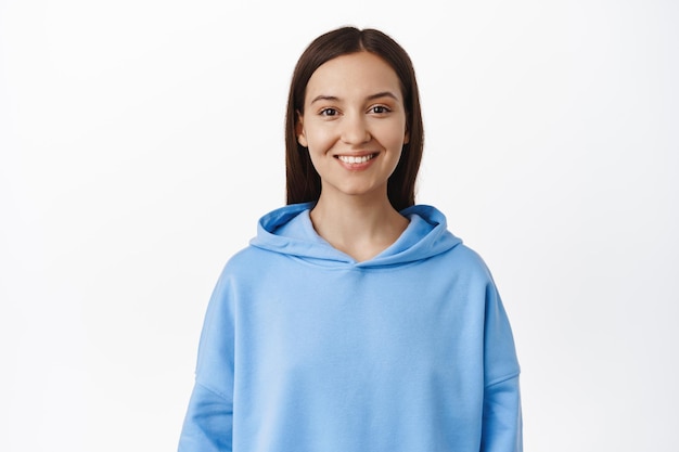 Portret młodej brunetki w wieku 20 lat, ubrana w niebieską bluzę z kapturem, uśmiechnięte białe zęby, wyglądająca na szczęśliwą, stojąca na białym tle.