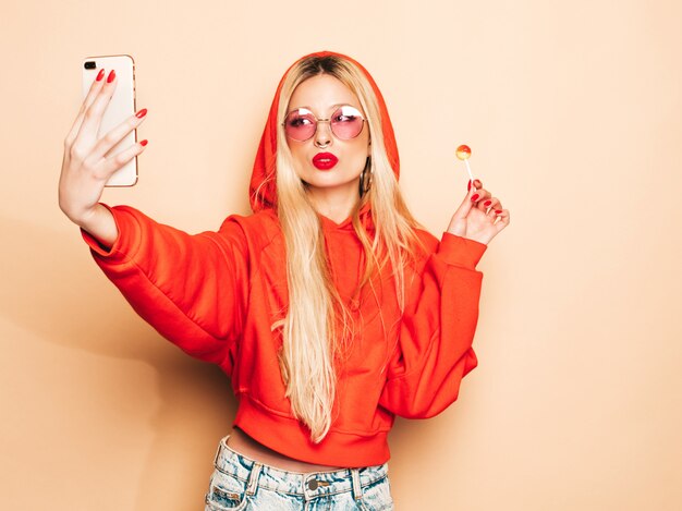 Portret młodej dziewczyny piękne hipster zły w modne dżinsy ubrania i kolczyk w nosie. Seksowny beztroski uśmiechający się blond kobieta bierze selfie. Model pozytywny lizanie okrągłe cukierki cukrowe