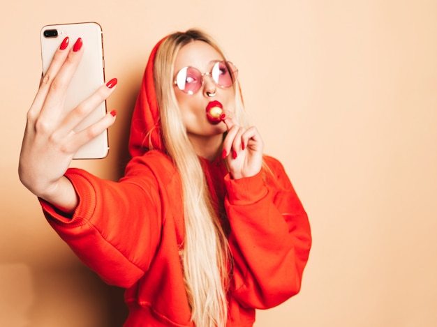 Portret młodej dziewczyny piękne hipster zły w modne dżinsy ubrania i kolczyk w nosie. Seksowny beztroski uśmiechający się blond kobieta bierze selfie. Model pozytywny lizanie okrągłe cukierki cukrowe