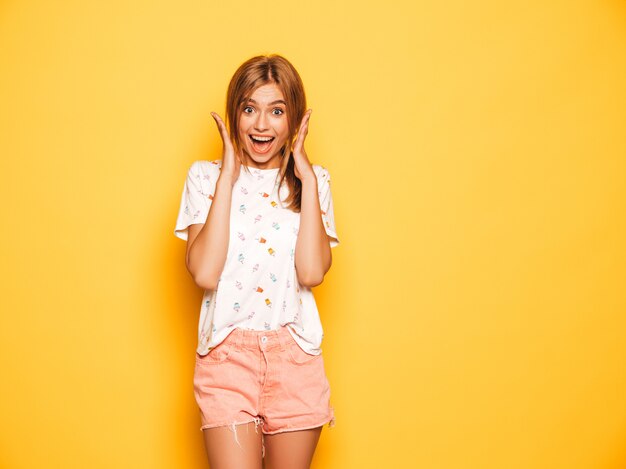 Portret młodej dziewczyny piękne uśmiechający się hipster w modne letnie dżinsy szorty ubrania. Seksowna beztroska kobieta pozuje blisko kolor żółty ściany. Pozytywny model zabawy. Wstrząśnięty i zaskoczony