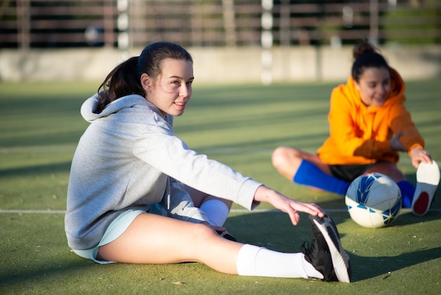 Portret młodej dziewczyny rozgrzewki przed grą w piłkę. Dwie piękne dziewczyny w strojach sportowych, siedząc na zielonym polu i rozciągając nogi, aby rozpocząć trening. Koncepcja zdrowego stylu życia i aktywności sportowej