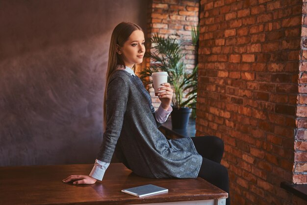 Portret młodej dziewczyny ubranej w elegancką szarą sukienkę trzyma filiżankę kawy na wynos odwracając wzrok siedząc na stole w pokoju z wnętrzem na poddaszu.