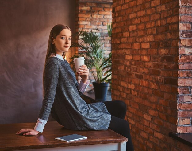 Portret młodej dziewczyny ubranej w elegancką szarą sukienkę trzyma filiżankę kawy na wynos patrząc w kamerę siedząc na stole w pokoju z wnętrzem na poddaszu.