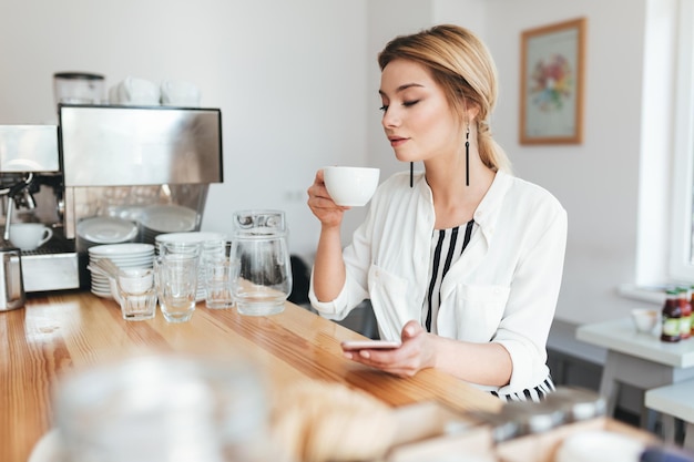 Portret młodej dziewczyny zamyślony z filiżanką kawy i telefonem komórkowym w ręce w kawiarni. piękna pani z blond włosami siedzi przy ladzie i pije kawę w kawiarni