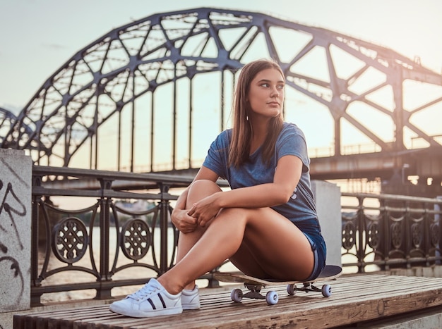 Portret Młodej Kobiety Brunetka Ubrana W T-shirt I Szorty, Siedząc Na Deskorolce Na Tle Starego Mostu O Zachodzie Słońca.