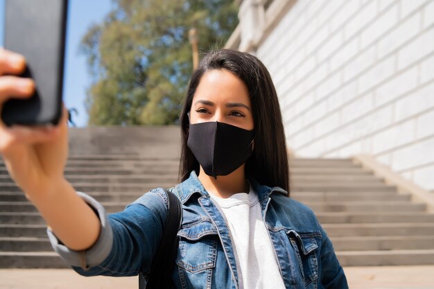 Portret młodej kobiety noszącej maskę i robienia selfie ze swoim telefonem mophile, stojąc na zewnątrz