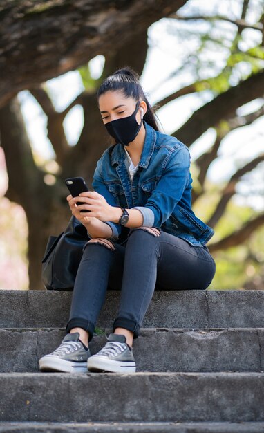 Portret Młodej Kobiety Noszącej Maskę I Używając Jej Telefonu Komórkowego, Siedząc Na Schodach Na Zewnątrz. Nowa Koncepcja Normalnego Stylu życia. Koncepcja Urbanistyczna.