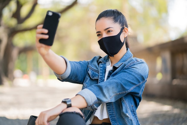 Portret młodej kobiety noszącej maskę na twarz i robienia selfie ze swoim telefonem mophile na zewnątrz