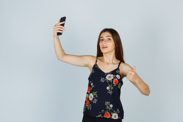 Portret młodej kobiety pokazując kciuk do góry podczas robienia selfie na smartfonie w bluzce i patrząc zadowolony widok z przodu