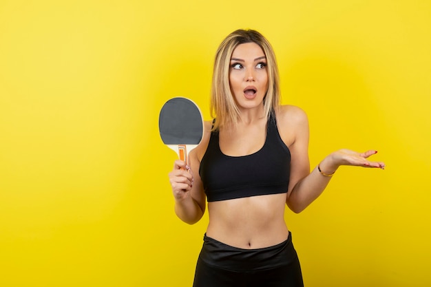 Portret młodej kobiety stojącej i trzymającej rakietę do tenisa stołowego na żółtej ścianie.