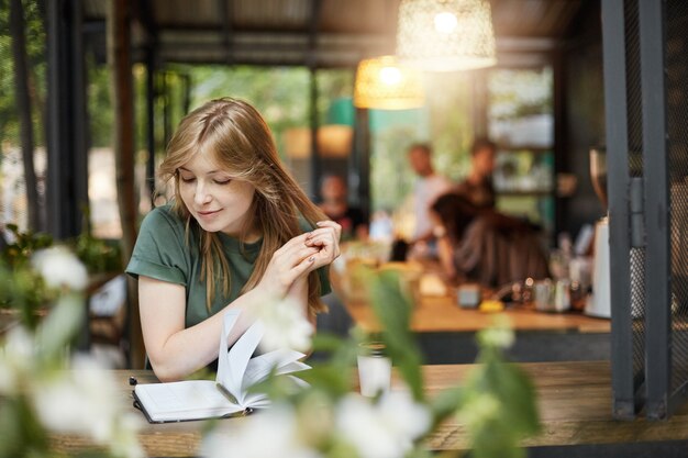 Portret młodej kobiety studentka blondynka czytająca notatki picia kawy w kawiarni na świeżym powietrzu, czekając na jej zajęcia z uśmiechem.