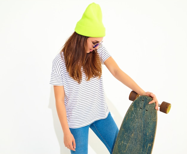 Bezpłatne zdjęcie portret młodej kobiety stylowe model w letnie ubrania w żółty czapka kapelusz pozowanie z biurkiem longboard. pojedynczo na białym