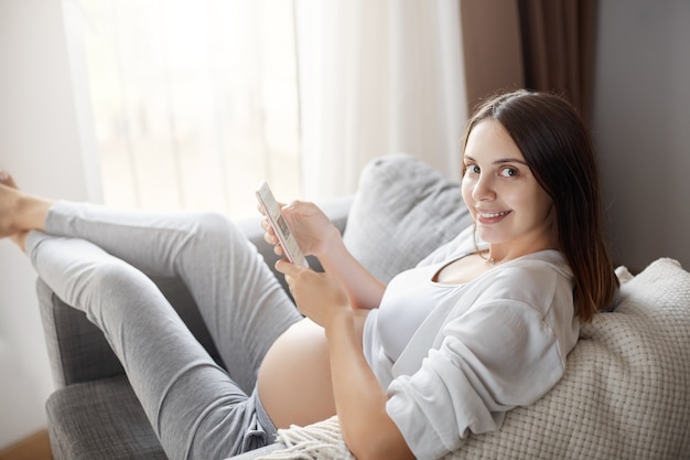 Portret młodej kobiety w ciąży pobyt w domu za pomocą inteligentnego telefonu. Czatuje z przyjaciółmi online.
