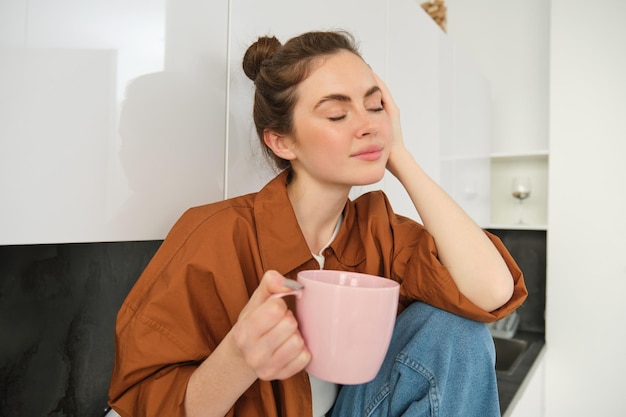 Portret młodej kobiety z filiżanką kawy siedzącej w kuchni i pijącej aromatyczny napój w domu