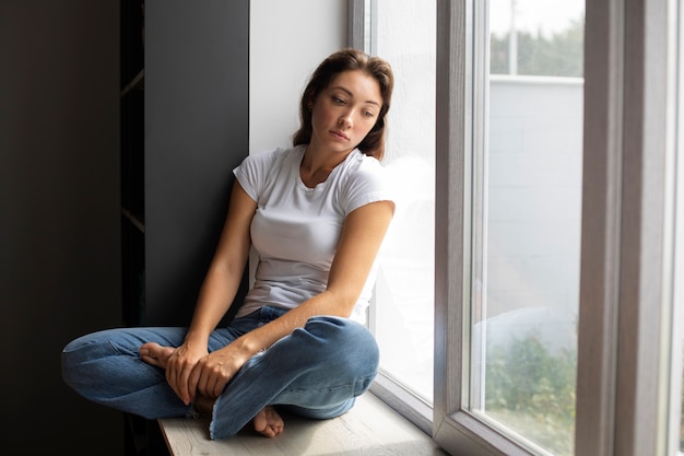 Bezpłatne zdjęcie portret młodej kobiety z niską samooceną siedzącej przy oknie w domu